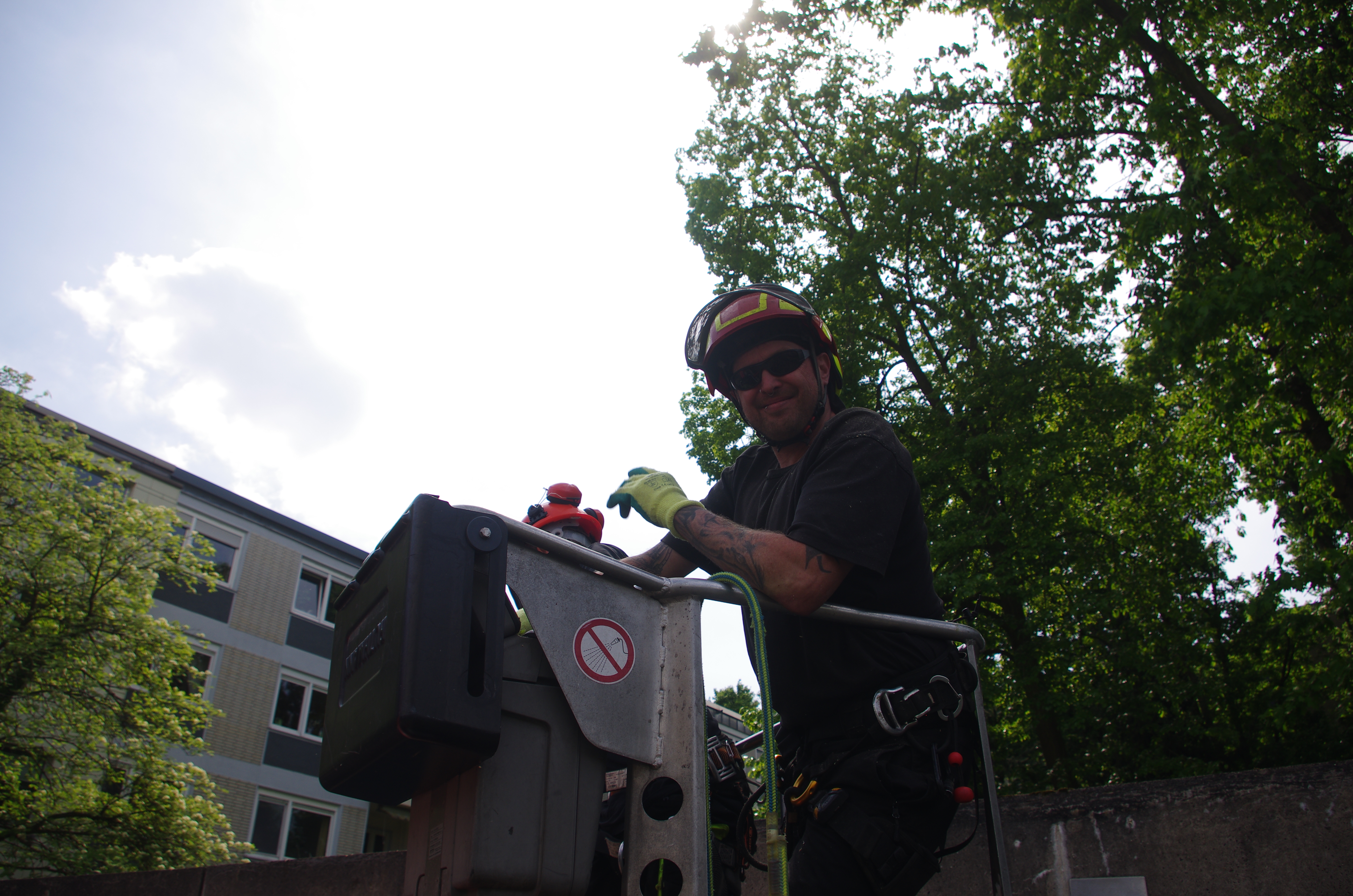 Baumpfleger mit Schutzhelm, Sonnenbrille und Arbeitshandschuhen bedient lächelnd eine Arbeitsbühne, im Hintergrund Baumkronen und ein Gebäude
